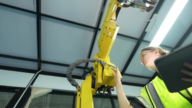 Low angle view of female engineer optimizing industrial automation. Professional woman using tablet to calibrate yellow robotic arm for future smart manufacturing efficiency.