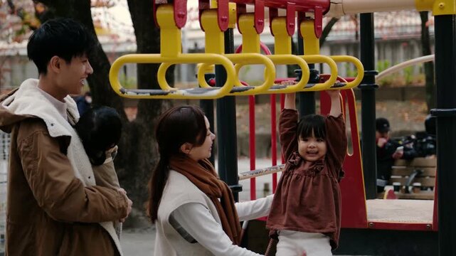 Young Japanese parents watch their daughter play on the monkey bars in a city park