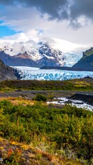 Naklejka premium Glacier view with green and yellow grass, mountains, and dramatic blue cloudy sky
