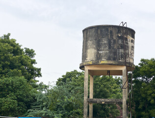 Elevated water storage tank with blue railings on stilts, standing against green trees and cloudy sky, a common rural water supply infrastructure.
