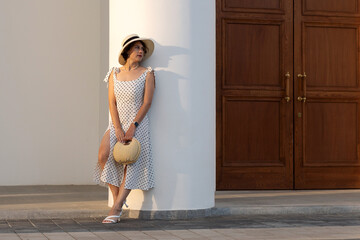 Woman in white polka dot dress and straw hat leans against white pavilion column holding woven circular handbag, bathed in golden hour light casting soft architectural shadows