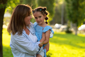 Still nestled in her mother's embrace, the young girl glances directly at the camera with a gentle smile, her hair tied in playful buns and sunlight highlighting her face