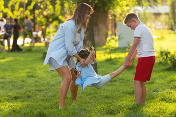 Mother and older brother whirl a little girl by her arms and legs in a sunlit city park, enjoying playful family time on lush grass during a warm afternoon