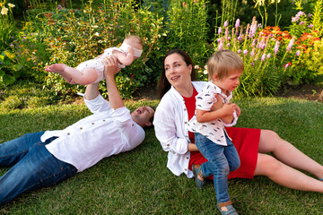 On the grass, father lies lifting the baby like airplane while mother holds their son, playful candid moment among blooming backyard flowers