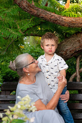 Grandmother embraces her young grandson while sitting on a bench under a large tree, sharing a tender moment in a lush garden setting