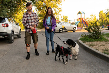 Man and his daughter walk two leashed dogs near parked cars and a colorful playground in a residential area during a calm evening stroll