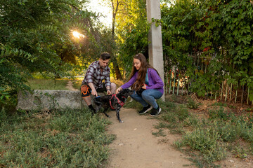 Father and his daughter kneel to pet one of their leashed dogs while walking outdoors, surrounded by greenery and warm evening sunlight filtering through the trees