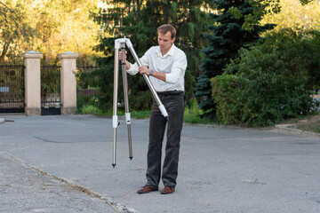 A man in formal casual clothing adjusts the legs of a tripod outdoors while preparing telescope equipment near stone structures and greenery