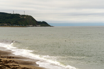 White waves break near the shoreline with a distant hillside topped by antennas and buildings, combining coastal water motion with landform and human presence. Sea of Okhotsk Sakhalin Island