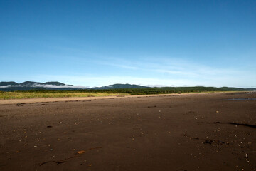 Vast sandy beach extends under clear blue sky, leading eye toward distant green hills and low mist clouds along coastal horizon in serene landscape. The coast of the Sea of Okhotsk, Sakhalin Island