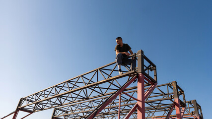 A man rests in a squatting pose at the top of a red steel frame structure during a break in his urban parkour routine under a wide open sky