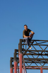 A male parkour athlete sits confidently atop an outdoor red steel parkour frame, feet on crossbars, with blue sky behind, demonstrating balance and body awareness