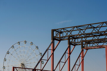 Ferris wheel rises in the background behind a red metal parkour structure under a clear blue sky, contrasting industrial geometry with amusement architecture in an open urban setting