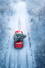 Aerial view of Christmas decorated car carrying Christmas tree on the road in a winter forest covered with snow in snowfall. Concept of Christmas transportation, shipping and delivery.