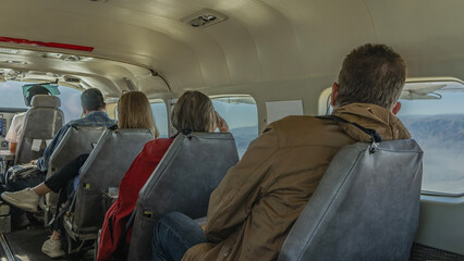 Passengers are sitting in a row on seats in a small plane. The pilot is ahead. View from the back....