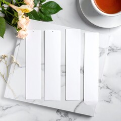 Four blank white bookmarks on a marble surface, accented with flowers and a teacup