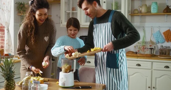 Family prepares fresh juice together in the kitchen, putting fruit into a blender. Healthy lifestyle, teamwork, and everyday family life at home.