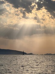 Sailboat on Calm SPeaceful seascape with a sailboat on calm water, distant mountains on the horizon and dramatic sun rays breaking through clouds. Golden sunset light reflecting on the sea surface. 