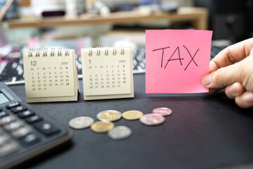 Hand holding TAX reminder note with coins and 2026 calendar for tax season and financial planning concept. tax season, financial burden, calculation of tax returns, and economic challenges.