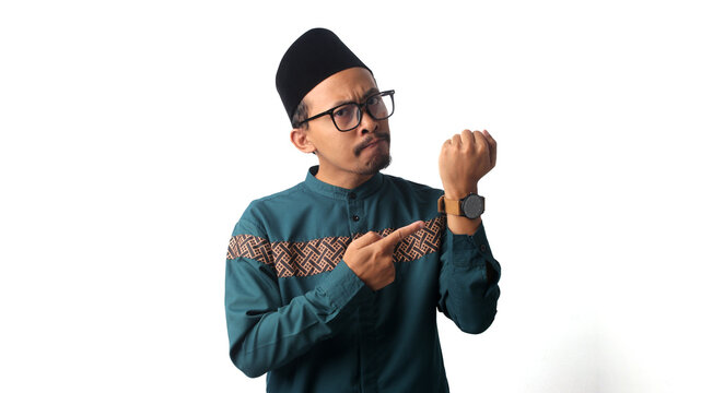 Impatient young Asian Muslim man pointing at his wristwatch while waiting for iftar, the time to break fasting during Ramadan. Isolated on a white background