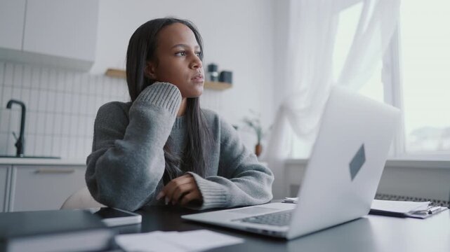 Freelance journalist or web-developer working in home workplace, female portrait . African american woman sitting at table with modern laptop and thinking, typing message and sending email to clients