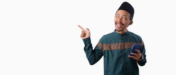 Asian Muslim man in a green traditional outfit holding a smartphone and pointing at empty copy space, isolated on white background. Perfect for promotional or communication themes