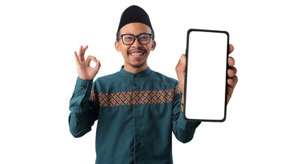 Asian Muslim man wearing a green traditional outfit and eyeglasses showing an OK hand gesture while presenting a smartphone with a blank white screen to the camera. Isolated on a white background