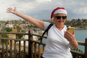 A middle-aged woman with a backpack on her back and wearing a Santa Claus hat is happy with the information she found on her phone against the backdrop of mountains, the old town and the sea.
