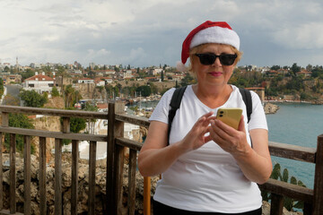 A middle-aged woman with a backpack on her back and wearing a Santa Claus hat is happy with the information she found on her phone against the backdrop of mountains, the old town and the sea.