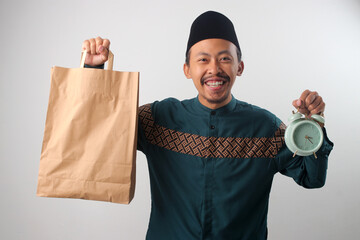 Asian Muslim man smiling at the camera while holding a brown paper bag and an alarm clock, Isolated on a white background. Food delivery, Ramadan preparation, or suhoor or iftar meal time concept