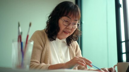 Smiling pretty senior asian woman painting and coloring on a canvas at home with creative lifestyle and healthy aging with mental wellness for retirement, elderly female enjoy leisure activity.
