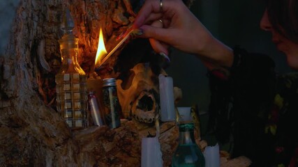 Close-up of woman lighting a candle on a ritual altar attached to a tree, surrounded by bones, bottles, and candles. Occult, witchcraft, or pagan ceremony at night. High quality 4k footage