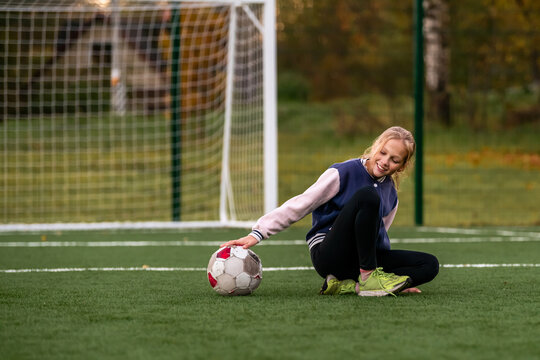 girl squats with a battered soccer ball on a green field covered