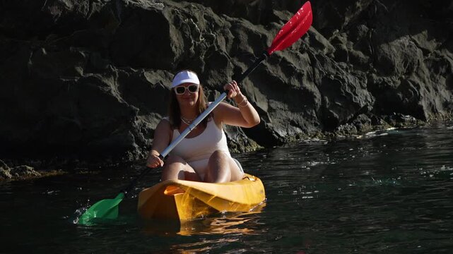 Kayak paddling woman enjoying sea travel along rocky cliff in bright sunshine