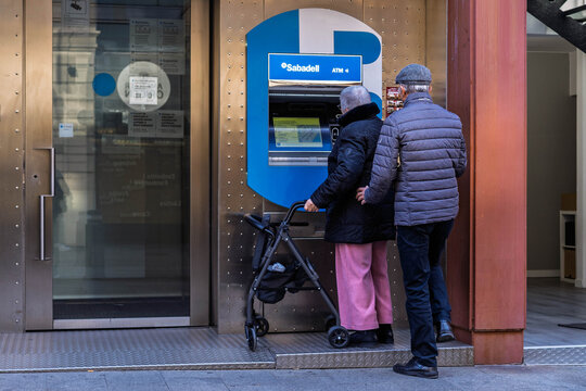 Barcelona, Spain-November 17, 2025. An older couple, one using a walker, withdrawing cash from a blue Banc Sabadell automated teller machine (ATM) on a city street.