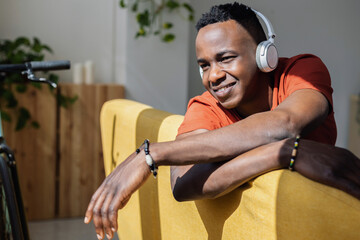Relaxed African man listening to music on sofa enjoying sunlight through window. Warm sunlight illuminates his face, creating a serene and cozy atmosphere of relaxation and mindfulness.
