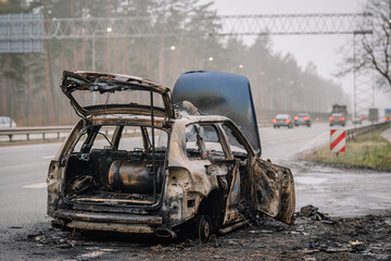 Burnt vehicle abandoned on roadside, with charred remains and smoke in the background, illustrating aftermath of fire incident and environmental impact