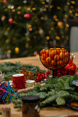 Festive table setting with a wire basket of fresh tangerines and Christmas decoration materials on a wooden surface. Blurred Christmas tree lights in the background create a cozy holiday atmosphere.