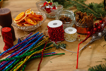 Variety of Christmas craft supplies on a wooden table, including dried orange slices, cinnamon sticks, colorful tinsel stems, and decorative beads. Creative DIY workshop and handmade materials.