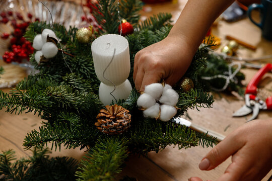 Close-up of woman's hands decorating a handmade Christmas floral arrangement with white cotton flowers, white candle, and pine cones in a cozy workshop.