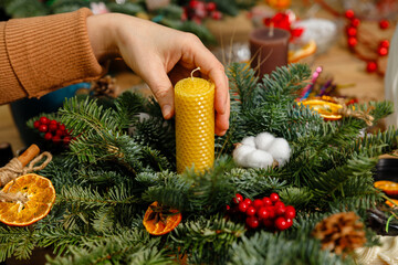 Close-up of a florist's hand placing a yellow honeycomb beeswax candle into a decorative Christmas wreath made of fresh fir branches, cotton, and dried oranges. Horizontal DIY workshop concept.
