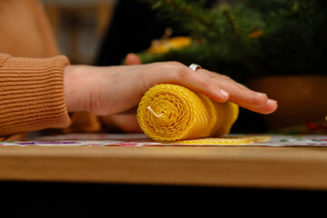 Close-up of a person's hands rolling a natural yellow beeswax sheet into a candle. Artisan craft process of making handmade traditional honey candles on a wooden table.