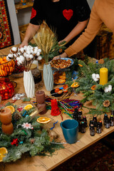 Two women hands creating handmade Christmas wreaths and centerpieces on a wooden table filled with festive decorations, candles, and essential oils. Creative workshop process DIY crafts concept.