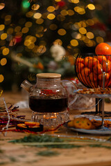 A cozy Christmas still life featuring a glass teapot on a candle warmer with tea, a basket of tangerines, and cookies on a wooden table against a blurred background of festive golden lights.