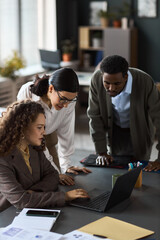 Young adult Caucasian woman sitting at desk using laptop while young adult Black man and young Caucasian woman standing beside her, observing screen in modern office setting © DragonImages