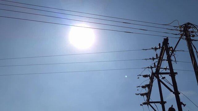 A bright sun and blue sky loom over complex power lines, a metal frame transformer, and utility poles. A typical rural electricity connection, emphasizing the interplay of light and infrastructure.