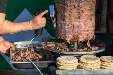 Close up of a chef shaving meat from a vertical shawarma grill with an electric knife, fresh pita breads stacked beside the rotisserie at an outdoor street cafe 