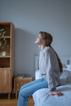 Mentally exhausted young woman sitting on bed with painful suffering expression. Tired female with signs of depression and fatigue sits in bedroom with closed eyes, struggling with inner problems.