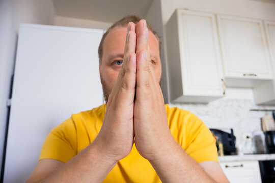 Adult man in a yellow shirt holds his hands together in a prayer or pleading gesture while looking at the camera in a home kitchen. Soft light with shallow focus.