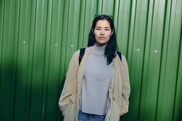 Woman wearing a beige coat and gray turtleneck stands before a green wall, casual street fashion, outdoor urban style, confident pose ideal for stock photos and editorial use